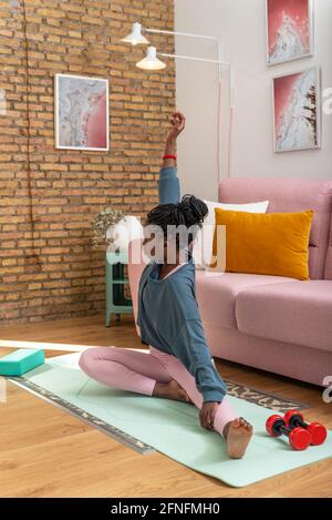 Black female in activewear stretching before workout at home while sitting on mat Stock Photo