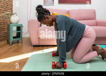 Side view of concentrated black sportswoman exercising with dumbbells on mat during workout at home Stock Photo