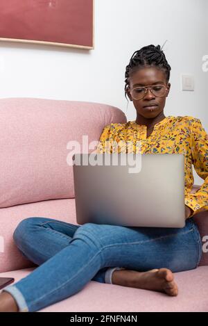 Focused African American female freelancer sitting on sofa at home and typing on netbook while working on project Stock Photo