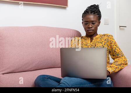 Focused African American female freelancer sitting on sofa at home and typing on netbook while working on project Stock Photo