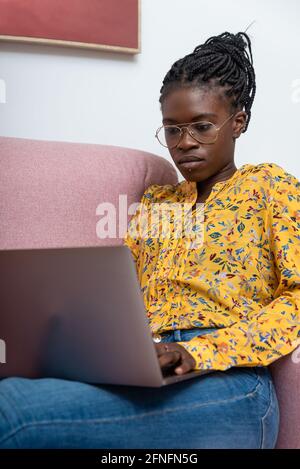 Focused African American female freelancer sitting on sofa at home and typing on netbook while working on project Stock Photo