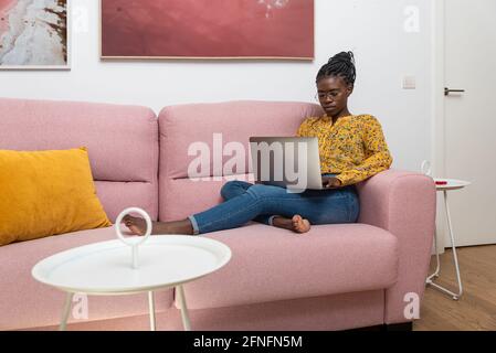 Focused African American female freelancer sitting on sofa at home and typing on netbook while working on project Stock Photo