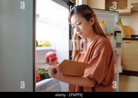 Young woman checking if she has enough fruits in fridge and making shopping list Stock Photo