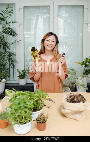 Lovely smiling young woman standing with hand trowel and gardening fork ready for plant repotting Stock Photo