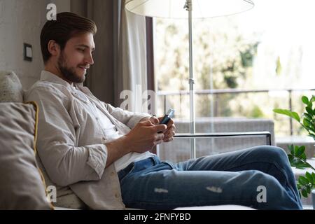 Handsome man in basic t-shirt smiling and holding mobile phone in hands while sitting on couch in living room. Stock Photo