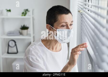 Senior patient in mask looking through the window while standing at hospital ward Stock Photo