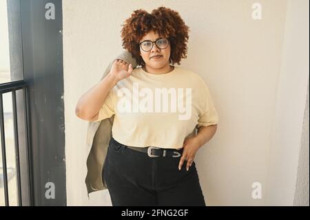 Beautiful happy smiling curvy plus size African black woman afro hair posing in beige t-shirt, jeans, stylish glasses on sunny balcony. Body imperfection, acceptance body positive diversity concept. Stock Photo