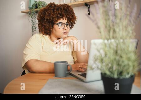 Attractive happy stylish plus size African black woman student afro hair in glasses studying online working on laptop computer at home office workspace. Diversity. Remote work, distance education Stock Photo