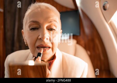 Mature woman applying lipstick near blurred mirror in plane Stock Photo
