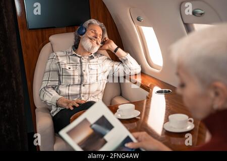 Smiling man in headphones sitting near wife reading magazine on blurred foreground Stock Photo