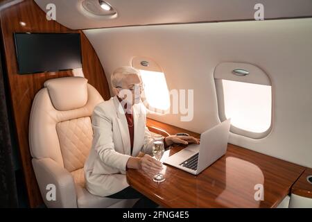 Mature businesswoman using laptop and holding glass of champagne in private plane Stock Photo