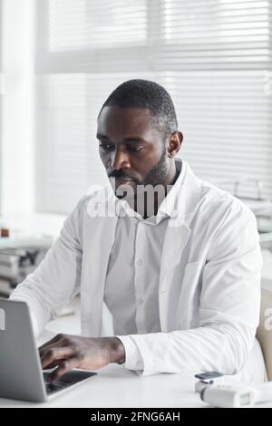 African young doctor in white coat sitting at the desk and working on laptop at his office Stock Photo
