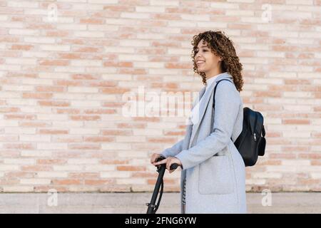 Content African American young female in casual wear riding scooter on paved sidewalk and looking away with smile Stock Photo
