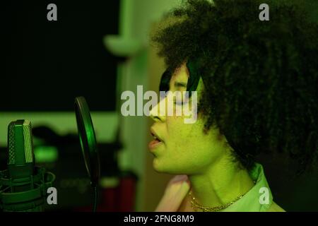 Black female singer performing song against microphone with pop filter while standing and closed eyes in sound studio Stock Photo