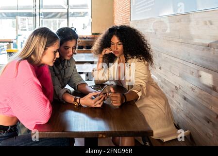 Young stylish female friends browsing mobile phones while sitting at table in cafeteria Stock Photo