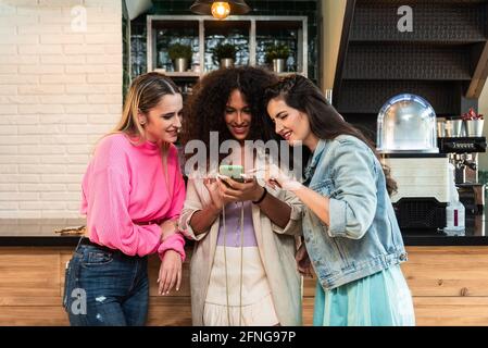 Young stylish female friends browsing mobile phone while standing in the cafeteria Stock Photo