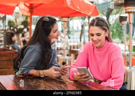 Optimistic young female friends in casual outfits using smartphones and discussing news while sitting at table on outdoor restaurant veranda on sunny Stock Photo