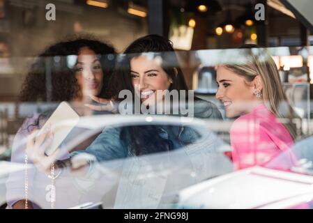 Through glass of cheerful young multiracial female best friends in trendy outfits smiling and showing two fingers gesture while taking selfie on smart Stock Photo