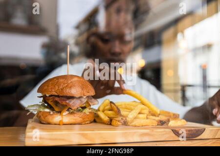 Through glass wall blurred African American female eating delicious fries and yummy hamburger served on wooden board on high table in fast food restau Stock Photo