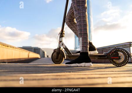 Cropped unrecognizable ethnic male entrepreneur riding electric scooter on city bridge walkway against buildings under cloudy blue sky Stock Photo