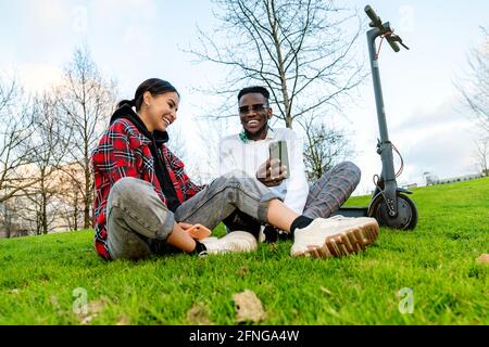 From below of cheerful multiracial couple in trendy wear with cellphone speaking on meadow with electric scooter in daytime Stock Photo