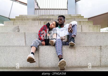 Young multiracial couple in trendy clothes sitting on city stairs with smartphone while looking away Stock Photo