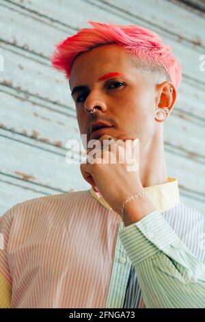 From below self confident thoughtful young gay in trendy apparel with pink hair and tattoos looking at camera Stock Photo
