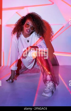 Full body attractive young African American female in stylish sunglasses sitting on floor and looking at camera in neon lights in dancing studio Stock Photo