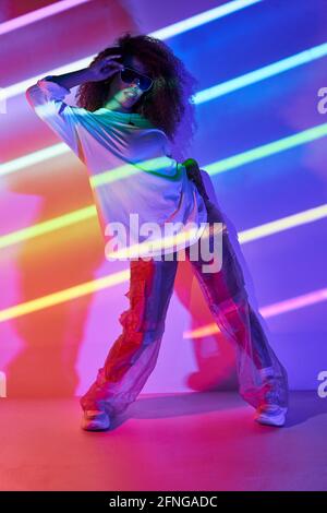 Full body stylish confident female dancer standing in neon lights and touching sunglasses in dancing studio Stock Photo
