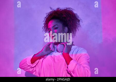 Portrait of joyful young African American female in white clothes wearing headphones on neck and looking at camera while standing in dark studio in ne Stock Photo