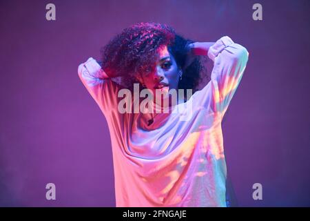 Portrait of young African American female dancer in loose informal wear touching curly hair and looking at camera while dancing in dark studio in neon Stock Photo