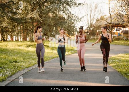 Smiling multiracial female runners in activewear jogging and talking during cardio training on walkway in town Stock Photo