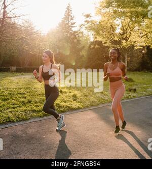 Smiling multiracial female runners in activewear jogging and talking during cardio training on walkway in town Stock Photo