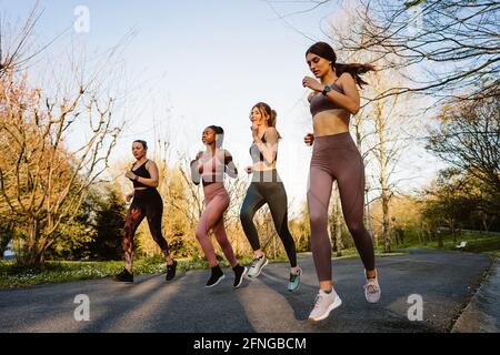 Smiling multiracial female runners in activewear jogging and talking during cardio training on walkway in town Stock Photo