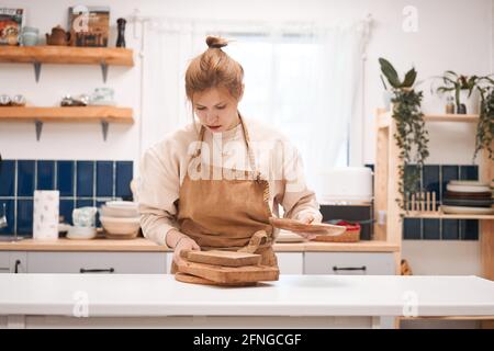 Young focused female in apron with assorted wooden chopping boards and plate at table in light house Stock Photo