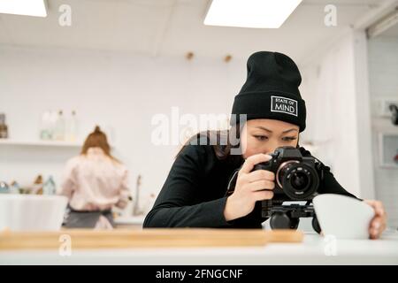 Ethnic female in beanie taking photo of bowl on digital camera against unrecognizable partner in kitchen Stock Photo
