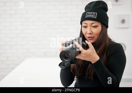 Ethnic female in black beanie and shirt using digital photo camera on blurred background Stock Photo