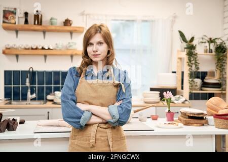 Young self assured female with folded arms in apron looking at camera at home Stock Photo