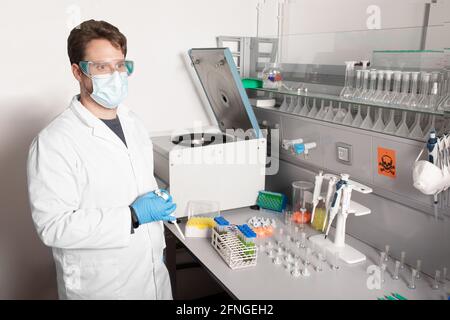 Male chemist in uniform and disposable mask looking forward at table with professional equipment and test tubes in laboratory Stock Photo