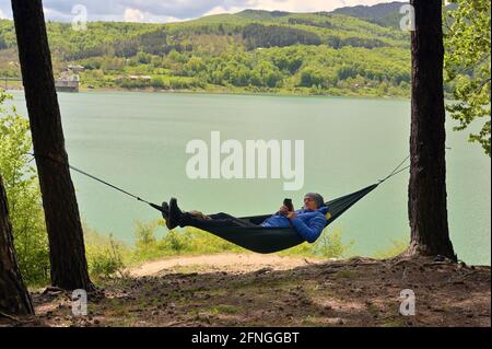 Young Man Using Mobile Phone While Relaxing In Hammock near Lake Stock Photo