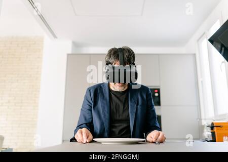 Stylish adult male pretending to eat in a modern kitchen experiencing virtual reality on the empty plate. Virtual reality glasses. Virtual food, Virtu Stock Photo