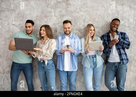 Group of diverse young friends with different gadgets studying or working together against grey studio wall Stock Photo
