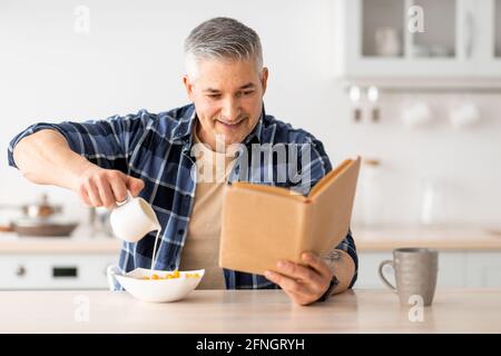 Happy senior man reading book and poring milk in cereals, having breakfast in kitchen interior Stock Photo