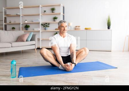 Active mature man sitting in butterfly asana , doing yoga on sports mat at home in living room, empty space Stock Photo