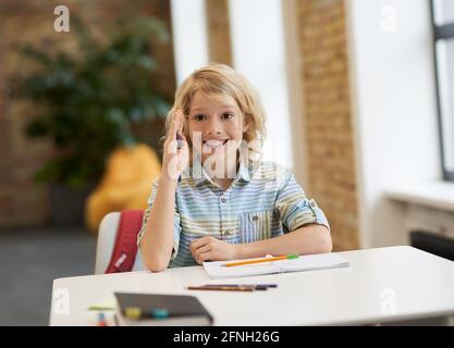 Energetic student. Motivated school boy smiling away raised his hand while sitting at the desk in classroom Stock Photo