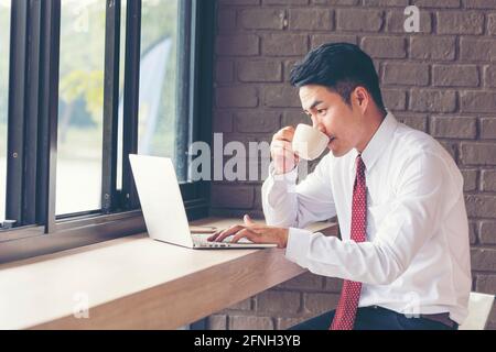Asian Handsome businessman working outdoor in the coffee shop with laptop and holding cup of coffee on hand. Work outside office using technology with Stock Photo