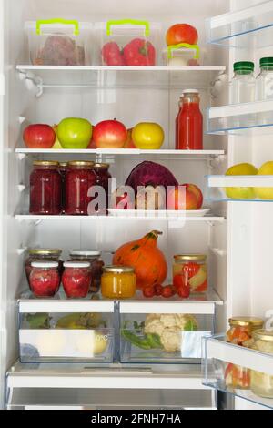 Variety glass jars homemade fruit and berry jams, juices and fresh vegetables and fruits on shelf in fridge. Fermented healthy vegetarian food. Stock Photo