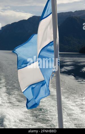 Blue and white Argentine national flag on mast in boat with water trail and mountains in the background in Lake Nahuel Huapi Stock Photo