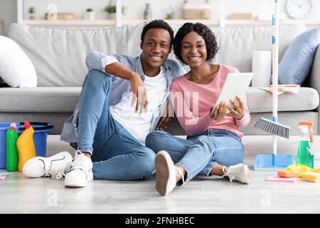 Black couple with cleaning tools resting, using digital tablet Stock Photo