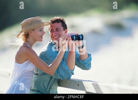 Lifestyle. Young couple with binoculars outdoors by wooden fence. Stock Photo